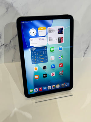 A black Apple iPad tablet standing upright on a clear acrylic display stand, showing the home screen with various app icons against a light marble-patterned background.