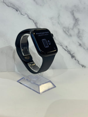A black Apple Watch with a black sport band, displayed on a clear acrylic stand against a white marble-patterned background.