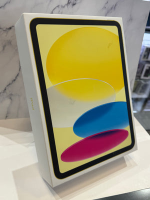 A white Apple iPad box standing upright on a white surface, with a colorful screen graphic showing yellow, blue, and pink abstract shapes. The background is a light marble-patterned wall.
