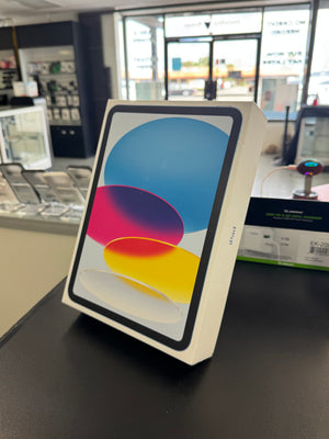 An Apple iPad in its original white box, standing on a black counter inside a retail store. The screen displays the colorful iOS home screen with blue, pink, purple, and yellow abstract shapes.