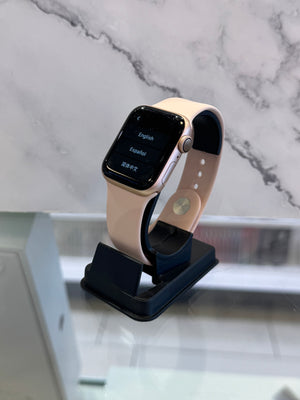 A smartwatch with a light beige band and a gold-toned case stands on a black charging dock against a white marble-patterned background.