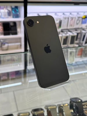 A black Apple iPhone shown from the back, resting on a clear display stand inside what appears to be a retail store with glass cases and electronic accessories in the background.
