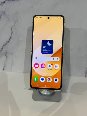 A smartphone with a black bezel and front camera cutout stands upright on a small metallic display stand against a light marble-patterned background.