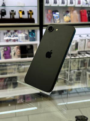 A black Apple iPhone shown from the back, resting on a clear acrylic display stand inside what appears to be a retail store or display case, with shelves of other phones and accessories blurred in the background.