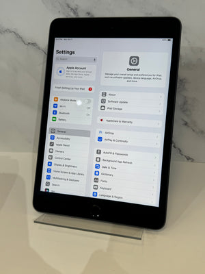 A black Apple iPad tablet on a clear acrylic stand, displaying the Settings menu on its screen against a white marble background.