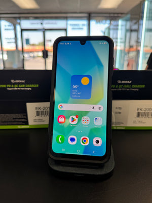 A black smartphone with a front-facing camera cutout stands upright on a charging dock on a dark tabletop. The screen is on and displaying colorful app icons and a weather widget.