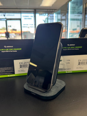 A black smartphone displayed upright on a gray charging dock, with two dark green and black Geekle PD & QC Car Charger boxes behind it in a retail or office setting.