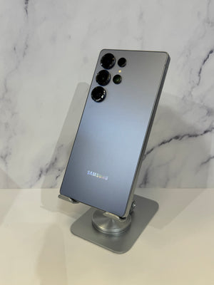 A Samsung smartphone shown from the back, resting on a metallic display stand against a white marble-patterned background.