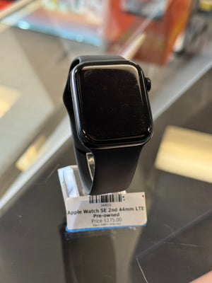 A black Apple Watch SE 2nd generation with a black band, resting on a dark reflective surface next to its retail price tag.