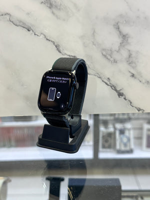 A black Apple Watch with a dark gray fabric band resting on a black charging stand, set against a white and gray marble-patterned background.