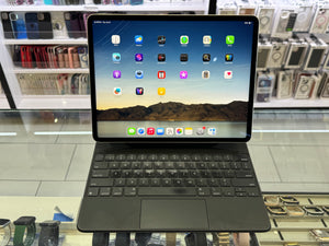 A black iPad with a matching keyboard case displayed on a glass counter in a retail store. The iPad screen is turned on and shows various app icons, with blurred shelves of electronics in the background.