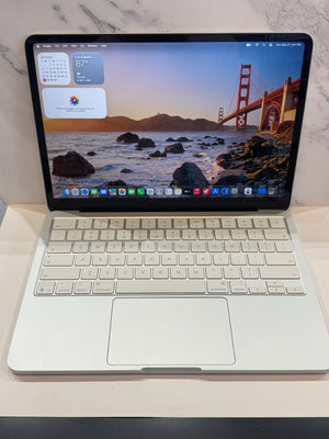 A silver aluminum laptop with a white keyboard and a large trackpad, shown open on a light surface against a white marble-patterned background. The screen is on and displaying a macOS desktop with a San Francisco Bay landscape wallpaper and the Golden Gate Bridge.