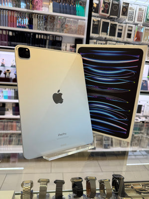 A silver Apple iPad Pro 2021 shown from the back, standing on a clear display stand next to a second iPad showing a colorful wallpaper. The setting is a retail store counter with various accessories in the background.