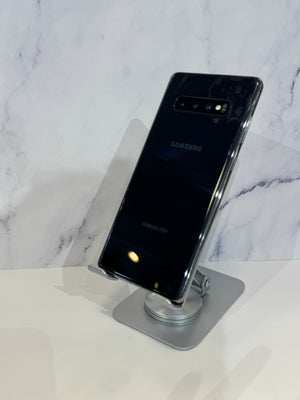 A black Samsung Galaxy S10+ smartphone shown from the back, resting on a clear acrylic stand with a silver base against a white marble-patterned background.