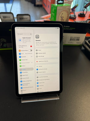 An Apple iPad standing upright on a clear display stand on a dark countertop, showing the Settings app screen. The background shows a retail or office environment with boxes and equipment.