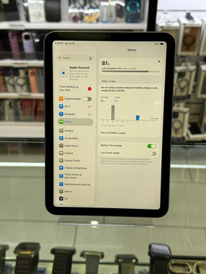 A black Apple iPad standing on a clear display stand in a retail store, showing the Settings app screen with battery-related options on the home screen.