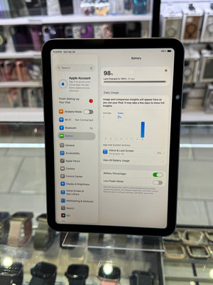 An iPad displaying the Settings app on its screen, resting on a clear display stand inside a retail store environment with glass cases in the background.