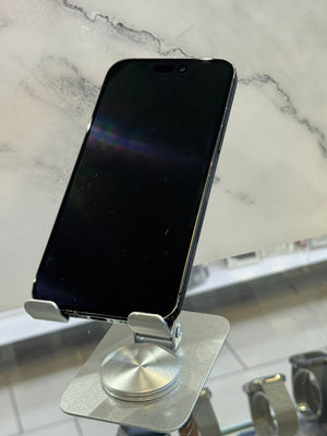 A silver metal magnetic phone mount with a square suction-style base and adjustable clamps, holding a black smartphone. The mount is shown attached to a glass surface on a white marble-patterned countertop.