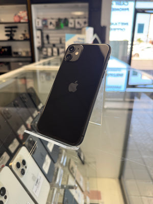 A black Apple iPhone 11 standing upright on a clear display stand inside a retail store. The back of the phone is visible, showing the Apple logo and dual camera system, with a glass display counter and other phones in the background.