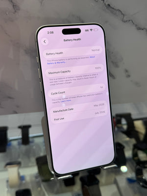 A smartphone displayed upright on a stand, showing the battery health screen on the home screen. The phone is photographed in a real retail or showroom environment with a marble-like wall and blurred display items in the background.