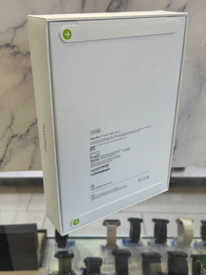 The back of a white Apple iPad Pro 13-inch M2 box, showing product specifications and regulatory text, displayed on a glass counter with a marble-patterned background.