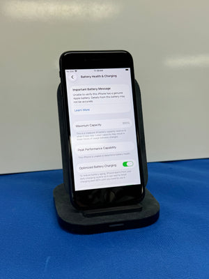 A black smartphone displayed upright on a black charging dock, showing a battery health and charging settings screen. The phone is photographed against a plain light background with a blue surface underneath.