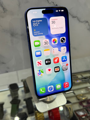 A blue iPhone with its screen turned on, displaying the home screen, resting on a white marble surface with a blurred store background.