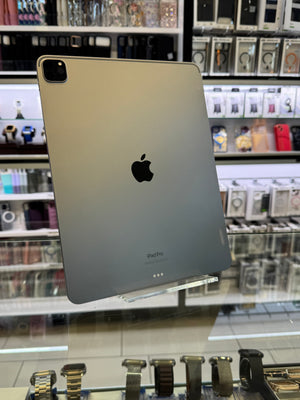 A silver Apple iPad Pro shown from the back, resting on a glass display counter inside a retail store with electronics and accessories visible in the background.