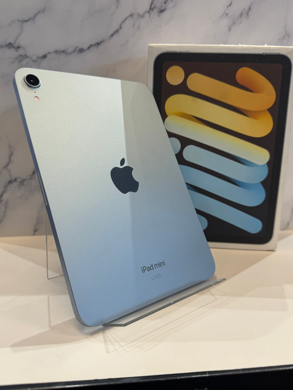 An Apple iPad mini in light blue-gray color, standing upright on a clear display stand with its retail box behind it against a marble-patterned wall.
