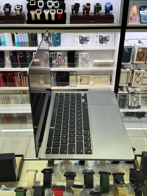 A silver aluminum laptop with a black keyboard and trackpad, shown open on a display stand inside a retail store with shelves of smartwatches and phone cases in the background.