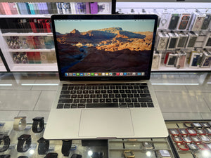 A silver Apple MacBook laptop with its screen on, displaying a desert canyon landscape wallpaper, set on a glass display counter in a retail store with watches and accessories in the background.
