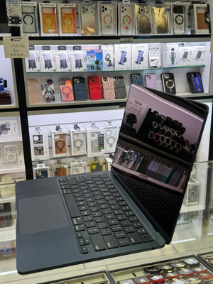 A dark slate blue laptop with a black keyboard and trackpad, shown open and angled in a retail store display with shelves of phone accessories in the background.