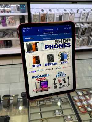 A smartphone with a dark red or black bumper case is shown sideways inside a retail electronics store, displaying a website on its screen.