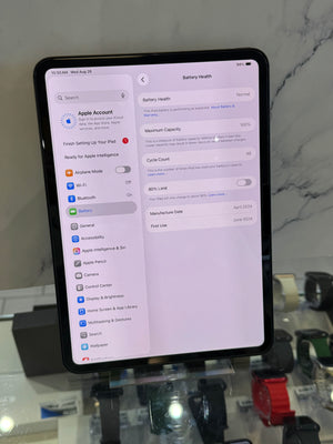 A black tablet displaying the Apple Settings app screen for Battery Health, standing on a reflective glass display counter in a retail store.