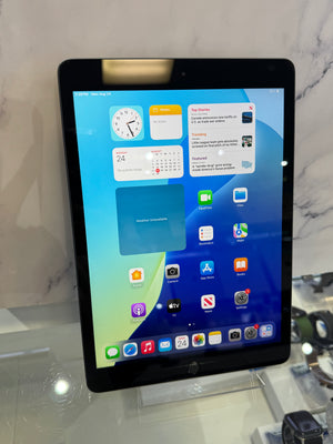 A black Apple iPad tablet on a clear display stand, showing the home screen with various app icons and widgets. The tablet is photographed indoors against a light marble-patterned wall and glass counter.
