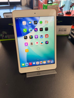 An Apple iPad tablet with a white front bezel and a gold-toned edge, standing on a clear acrylic display stand on a dark tabletop. The screen is on and displaying the iOS home screen with various app icons.