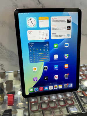 A tablet device with a black bezel and silver edges, displaying a blue and green home screen interface, photographed in a retail or showroom setting with glass display cases in the background.