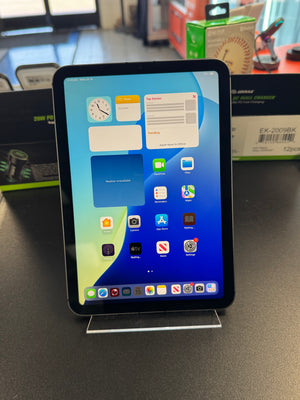 An iPad with its screen turned on and displaying the home screen, resting on a clear acrylic stand on a dark tabletop. The background shows some retail store clutter with boxes.