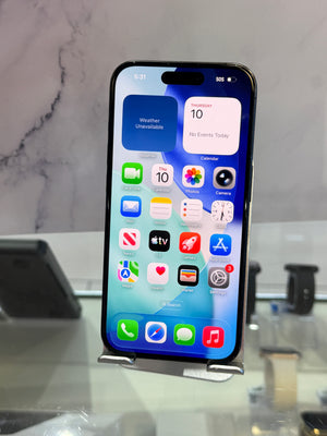 A black iPhone with a lit home screen displaying various colorful app icons, resting on a small metal display stand on a glass counter.