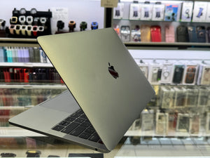 A silver Apple MacBook laptop with its lid open, standing on a glass display counter inside a retail store. The Apple logo is visible on the back of the lid, and shelves of other electronics and accessories are blurred in the background.