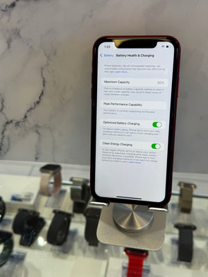 A smartphone in a dark case mounted on a display stand, showing the battery health and charging settings screen. The phone is photographed in a retail or showroom setting with a marble-like surface and other devices blurred in the background.