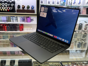 A dark gray Apple MacBook laptop with an open screen displaying a blue whale wallpaper, shown floating above a glass display counter in a retail store.