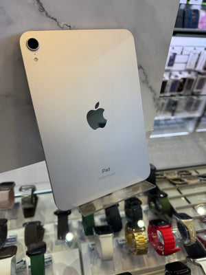 An Apple iPad shown from the back, featuring its silver aluminum finish and the iconic Apple logo, resting on a marble-patterned surface with a blurred watch store background.