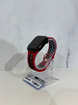 A smartwatch with a black rectangular face and a red perforated sport band, displayed on a clear acrylic stand with a price tag in front, against a white marble-patterned background.