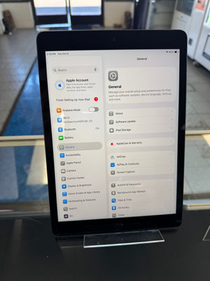 An iPad standing upright on a clear display stand on a dark tabletop, showing the Settings app on the screen in a retail or office environment.