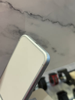 A close-up of a silver smartphone corner resting on a light marble-like surface, with a blurred display of other tech items in the background.