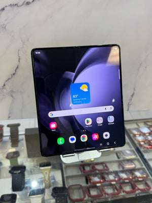 A black Samsung Galaxy Z Fold 3 smartphone displayed on a small stand in a retail setting, with a marble-patterned wall and watch display cases in the background.