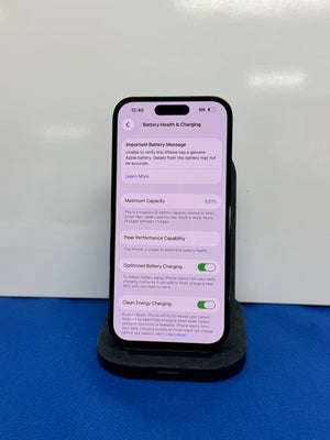 A black smartphone displayed upright on a black charging stand against a white wall, with a blue surface underneath. The phone screen shows battery health and charging settings.