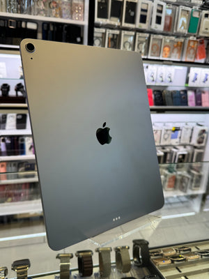 A silver Apple iPad shown from the back, featuring the iconic black Apple logo in the center. The tablet is photographed inside what appears to be a retail store, with display cases and electronic accessories blurred in the background.