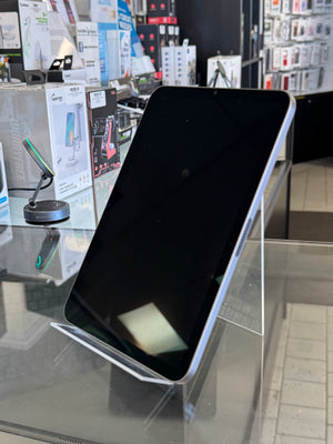 A black tablet or e-reader device with rounded corners displayed on a clear acrylic stand inside a retail store. The screen is off and black, and the background shows store fixtures, display boxes, and shelving.